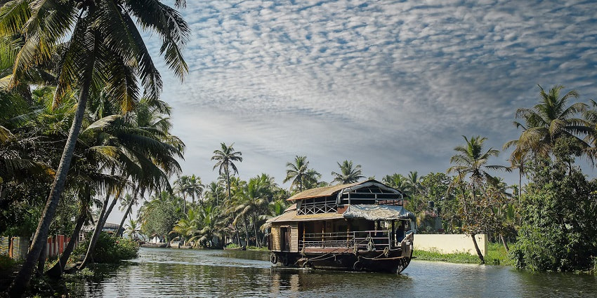 An alluring view of a houseboat cruising on the backwaters of Alappuzha surrounded by tall trees.
