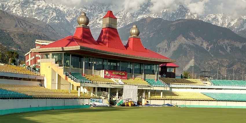 Dharamshala Cricket Stadium with vibrant green field and snow-capped mountains in the background.