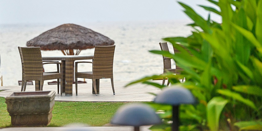 A stunning view of a restaurant near a sparkling beach with brown chairs and tables.