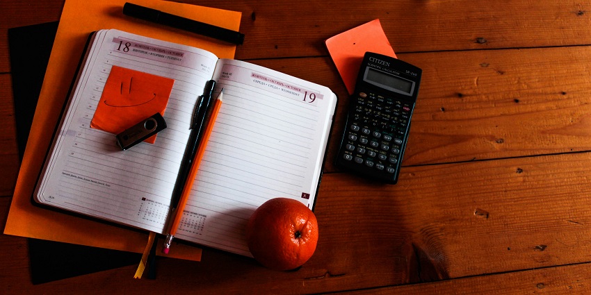 An aerial view of a notebook on a table with an orange kept on the side with a calculator.