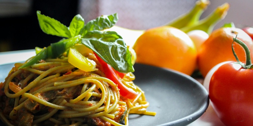Pasta served on a plate garnished with leaves and bell pepper with fruits on the side.