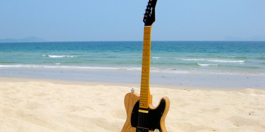 A guitar thrust in the sand on a beach with clear blue waves in the background.