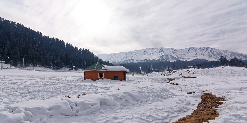 A landscape of Gulmarg with a snow-covered field, a cosy cabin, and majestic mountains