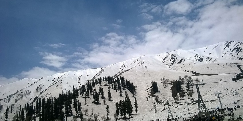 View of the snow capped hills in Gulmarg. There are green trees in the middle of the view.