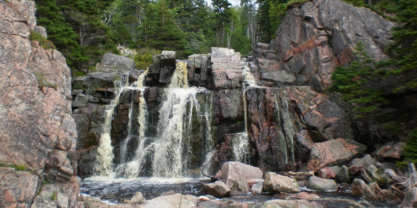 A picture of a waterfall with water cascading down a rocky mountain with trees and greenery nearby