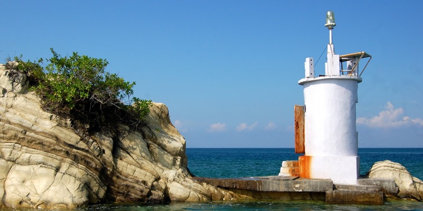 Havelock lighthouse in the middle of the beach with big rocks and bushes on both sides.