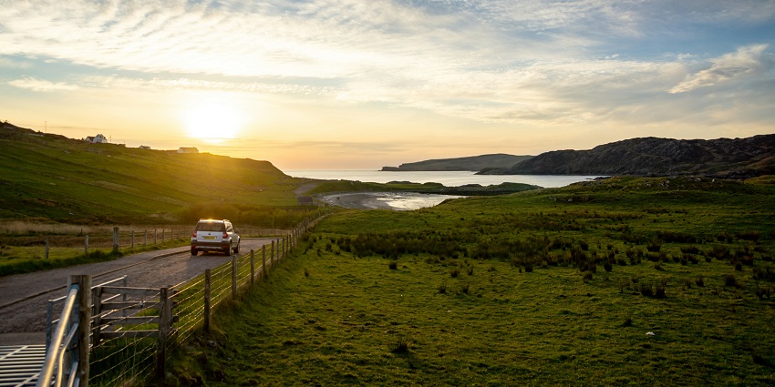 A view of a white car on the road in the middle of a green grassland with a lake in front.