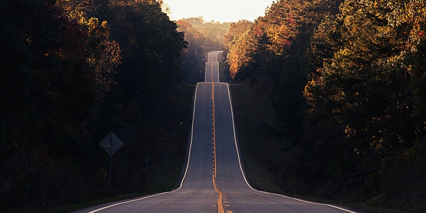 A distant view of a long road surrounded by lush green trees leading up to a hill.