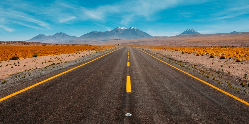 A view of a long road leading up to snow-covered mountains under a clear blue sky.