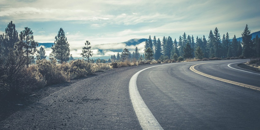 A view of a concrete road leading up to a hill encircled by lush green trees and clouds.