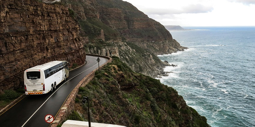 A distant view of a bus on a narrow road near a cliff surrounded by azure oceans.