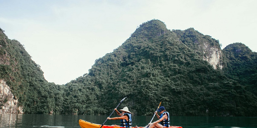 A frontal view of two people on an orange kayak on a water body against lush greenery.
