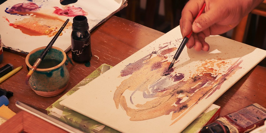 A person holding a paintbrush in hand and painting on a canvas with different colours.