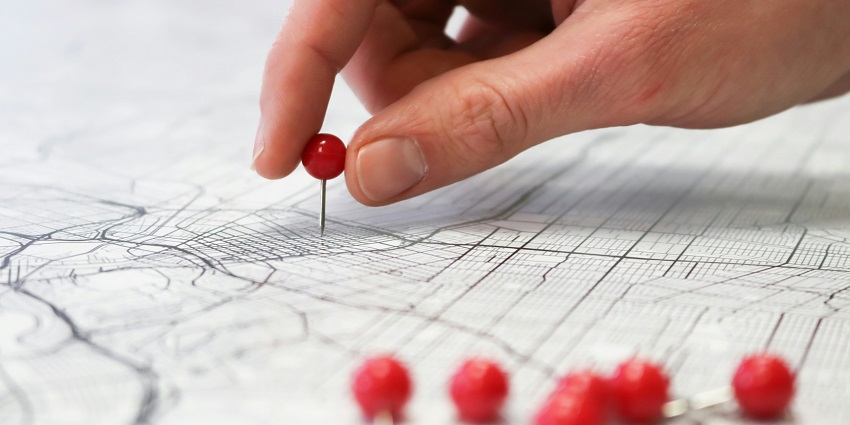 A close-up landscape of a map with a person holding a red pin to locate a place on it.