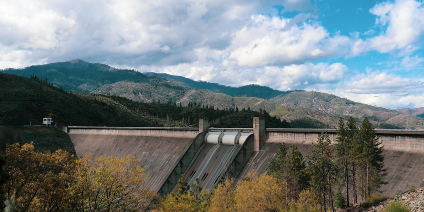 A wide view of a dam with soft clouds, lush greenery and mountains surrounding it.