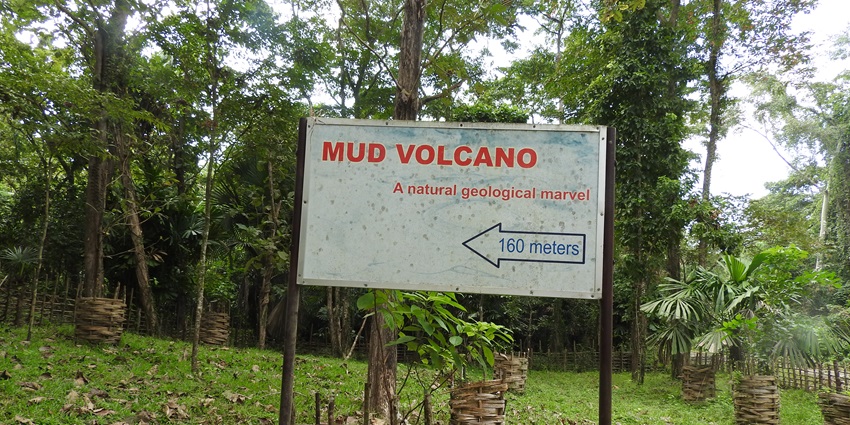 A white sign board with mud volcano written on it in the middle of a green forest.