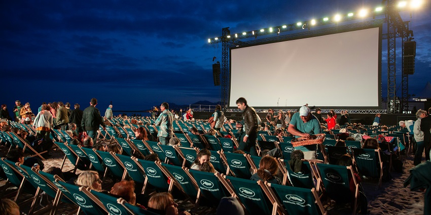 People gather on a beach at night, seated on chairs, waiting for a film to be screened.