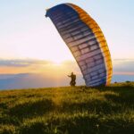 A silhouette of a person standing on lush green grass holding a paragliding parafoil.