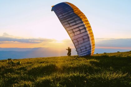 A silhouette of a person standing on lush green grass holding a paragliding parafoil.