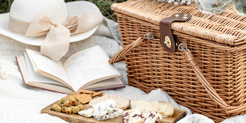 A mesmerising view of a brown picnic basket around an open book on a white bedsheet.
