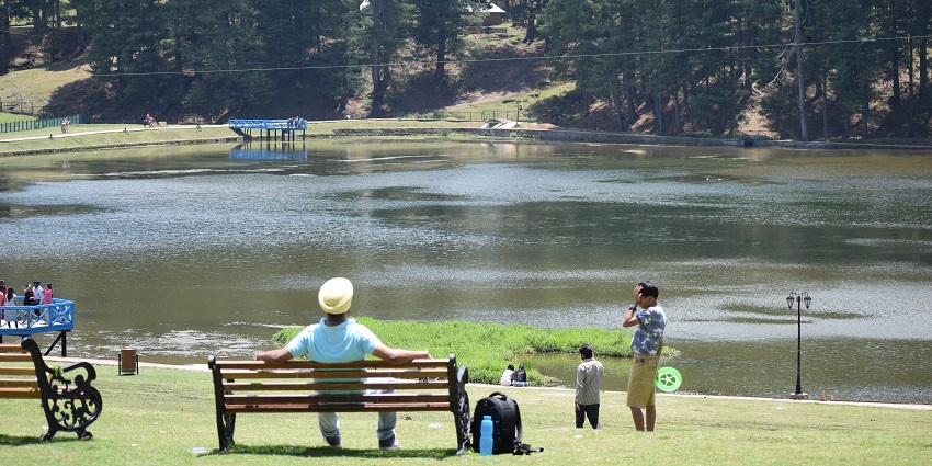 Serene Sanasar Lake surrounded by lush green hills and dense forest under a blue sky.