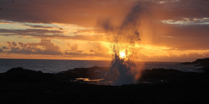 The serene view of a sea wave crashing on shore forming a sprouting blowhole at sunset.