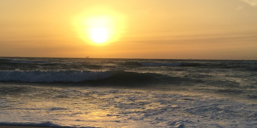 A fascinating view of waves crashing on a beach during sunset with a building far away.