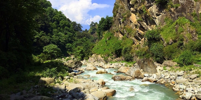 Tirthan River caseding through rocks, lush green mountains surrounded and clear blue sky.