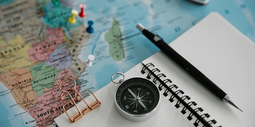A pen placed over a notebook that is on top of a map with places marked by a thumb pin.