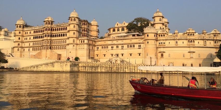A boat on a body of water with the backdrop of a palace in Udaipur, the City of Lakes