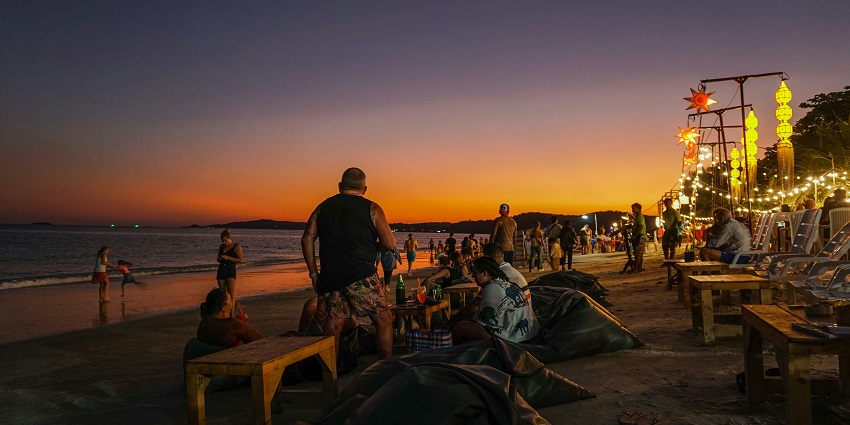 Lively beach gathering at sunset with people relaxing on beanbags, lights glowing warmly