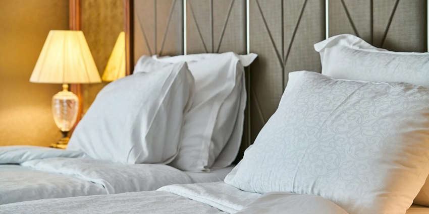 A room with two single beds with white bed sheets, pillows and a lamp on the side table.