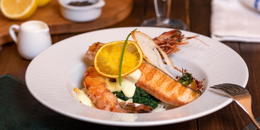 A white plate with a dish made with fish served on it and garnished with a sliced orange.