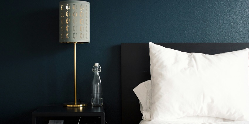 A view of a hotel room with white pillows and a grey lamp on the table beside the bed.