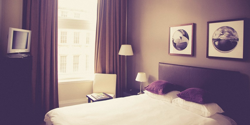 A view of a hotel room with a bed with white sheets, curtains, a television and a lamp.