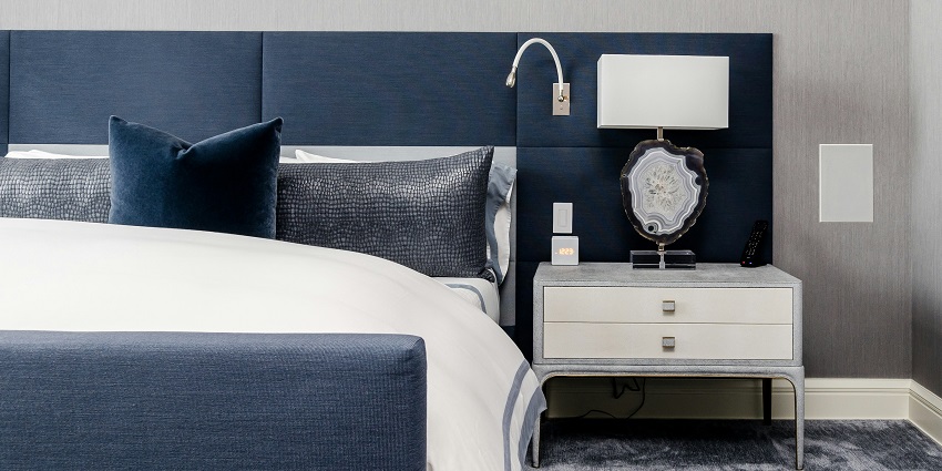 A room view with a bed with white sheets, blue pillows and a white table on the side.