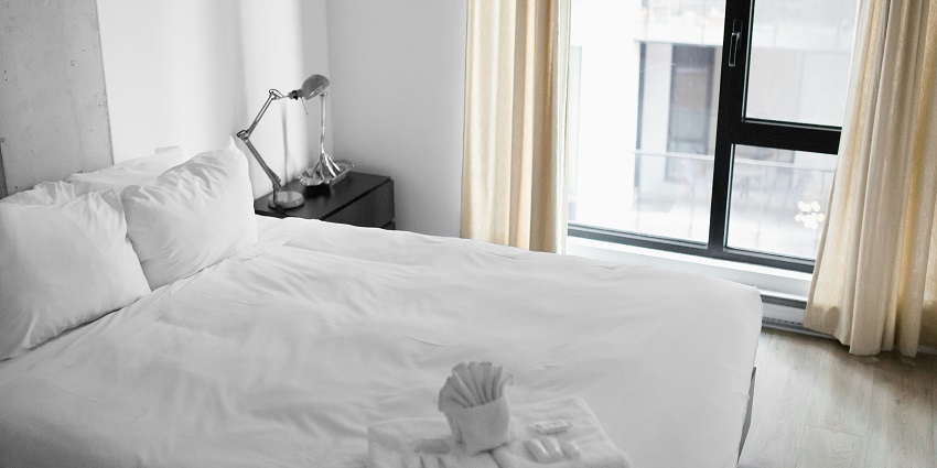 View of a hotel room with a white-lined bed, side table and open curtains during the day.