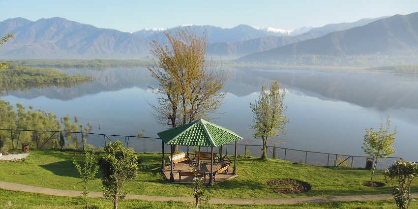 Wular Lake, surrounded by lush greenery and distant mountains under a clear blue sky.