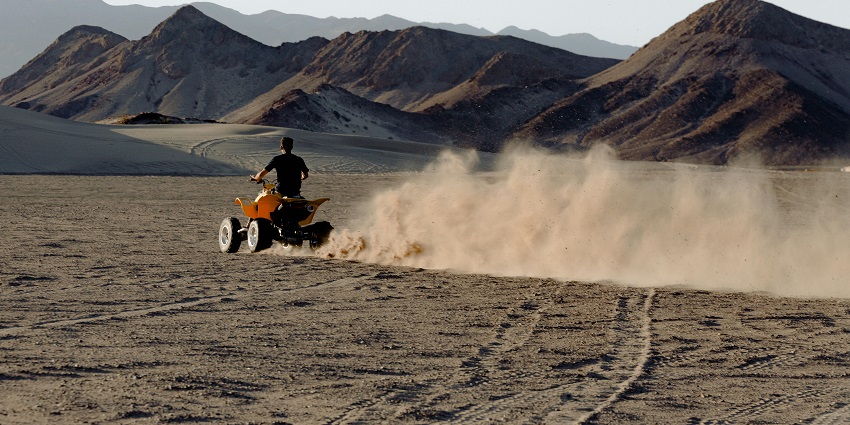 A man riding a dirt bike in a desert and leaving behind a ray of sand from its tyres