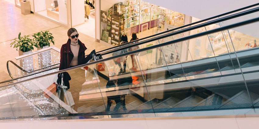 A picture of a woman on an escalator with shades on eyes and paper bags in both hands.