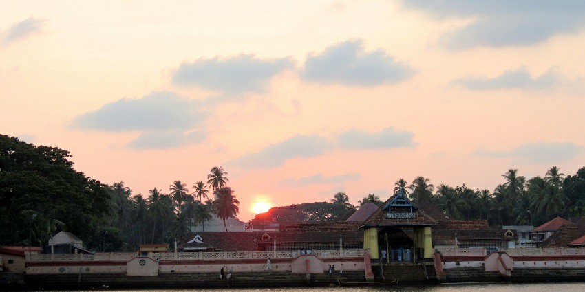 A distant view of a Thrissur temple from across a river with the sun setting