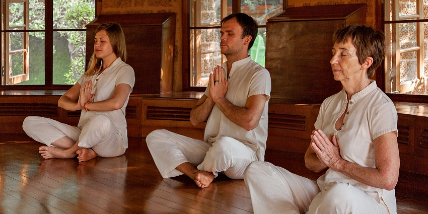A picture of two women and a man in white clothes practising yoga and meditation.