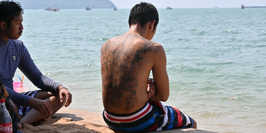 A man with a big tattoo on his back sitting at the beach, admiring the scenic views of the ocean