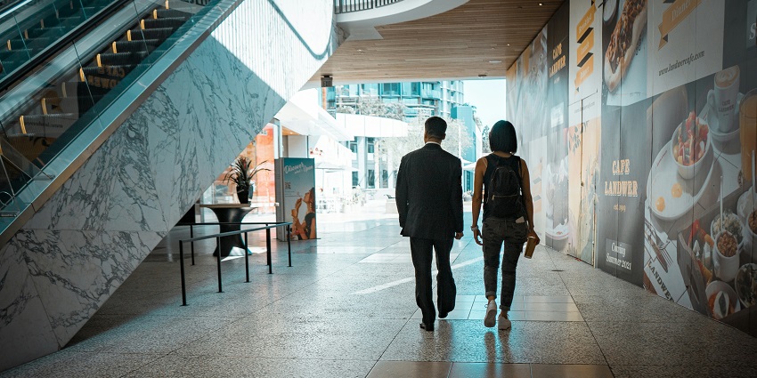 View of a man and woman walking in a hallway with an escalator on the left-hand side.