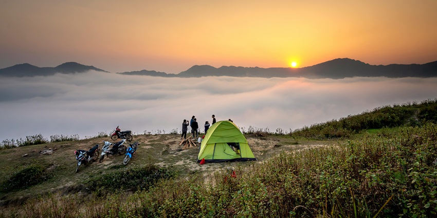 Group of people standing beside a green tent in a forested area with trees and sunlight -camping in Guwahati.