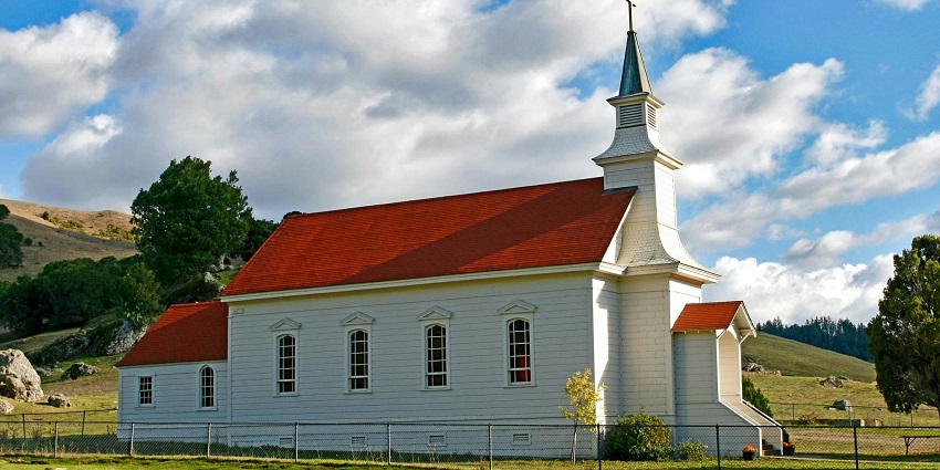 A red and white church stands on a green grass field under a clear blue sky.