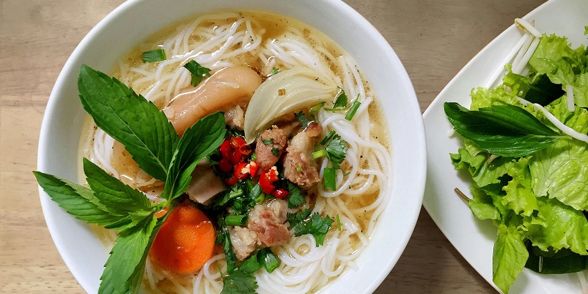 Image of a close-up of a bowl of cooked noodles, garnished with green herbs and spices.