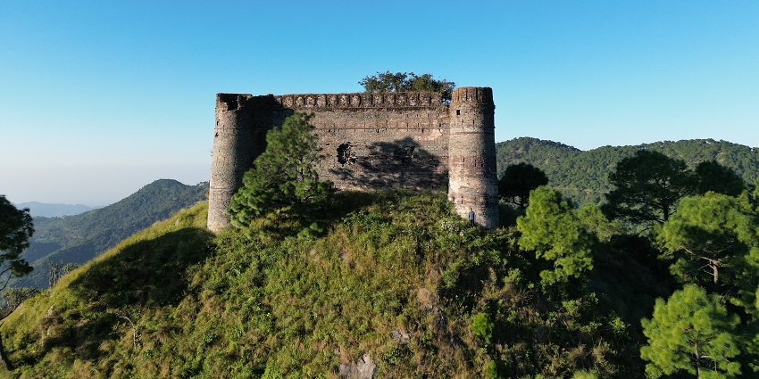 View of an old building on the top of a hill, surrounded by lush greenery & mountains.