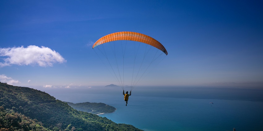 An image of a person paragliding in Thailand in the clear blue sky and sea behind.