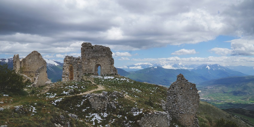 Image of stone ruins under a cloudy sky with weathered walls and overgrown vegetation.